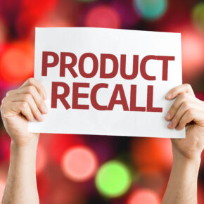 Hands holding product recall printed on a white paper with blurry lights in the background.
