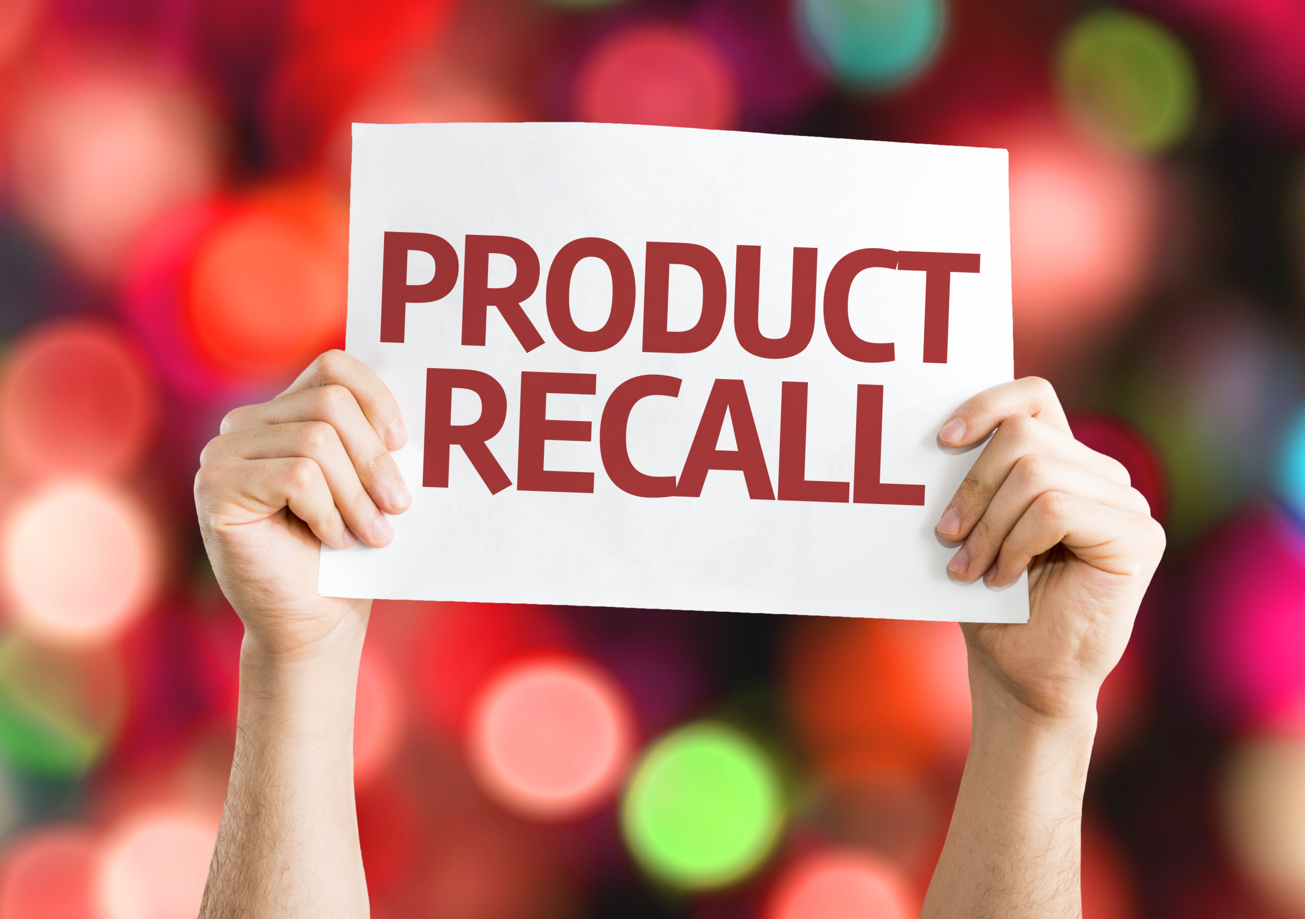 Hands holding product recall printed on a white paper with blurry lights in the background.