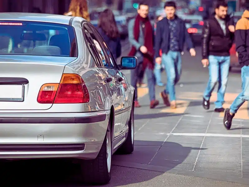 rear side of a silver car parked near a sidewalk. Several people are walking on the sidewalk beside the car, and the scene appears to be in an urban area with a paved street and pedestrian pathway.